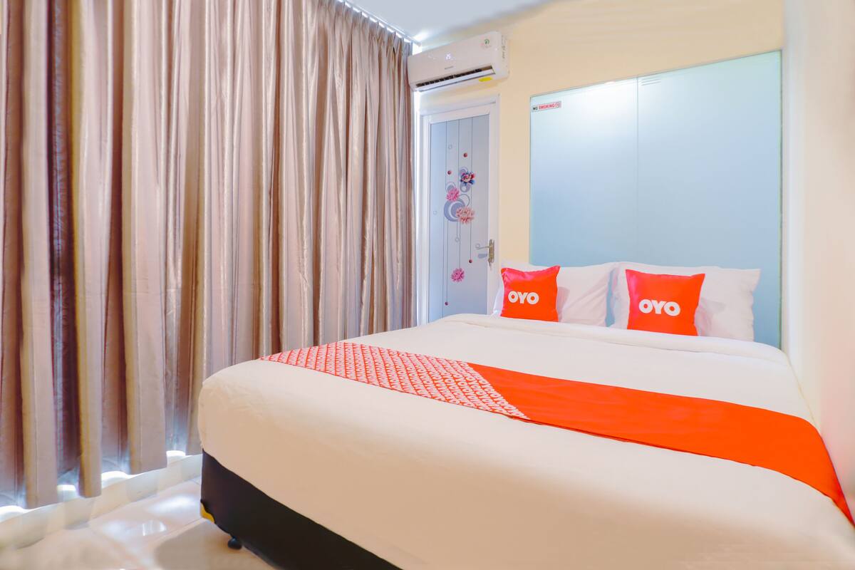 A hotel room with a large bed, two pillows with the brand name "OYO", a window with curtains, and an air conditioning unit on the wall.