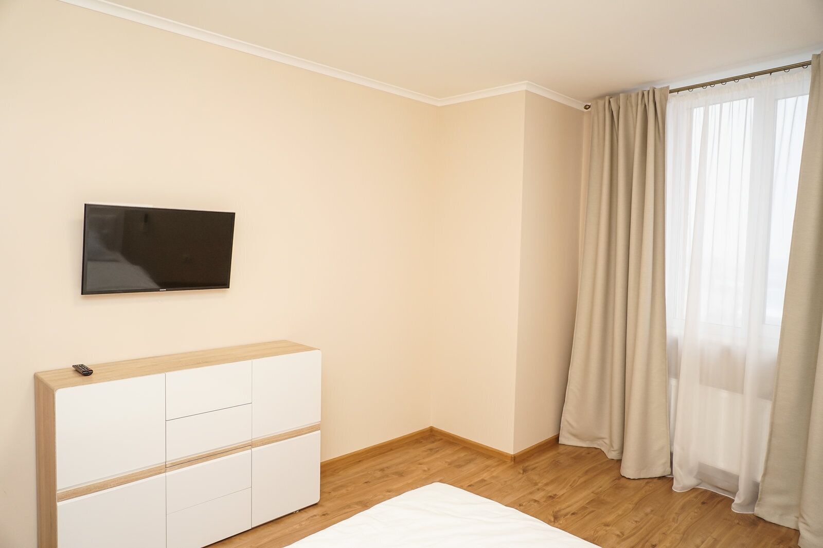 A bedroom with a bed, a TV mounted on the wall, a white dresser, and a window with sheer curtains.