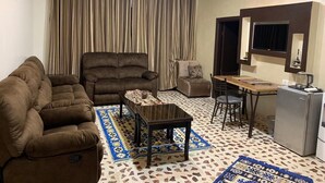 A living room with a sofa, armchair, coffee table, and a dining area with a table and chairs. There is a refrigerator and a window with curtains. - Mesha Stele Hotel Apartments (Madaba)