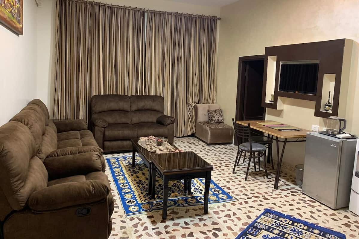 Ein Wohnzimmer mit einer Couch, einem Sessel, einem Couchtisch und einem Essbereich mit Tisch und Stühlen. Ein Kühlschrank und ein Fenster mit Vorhängen sind ebenfalls vorhanden.
