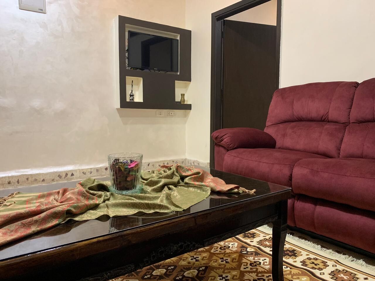 A living room with a dark brown sofa, a glass coffee table with a green cloth, and a wall-mounted TV.
