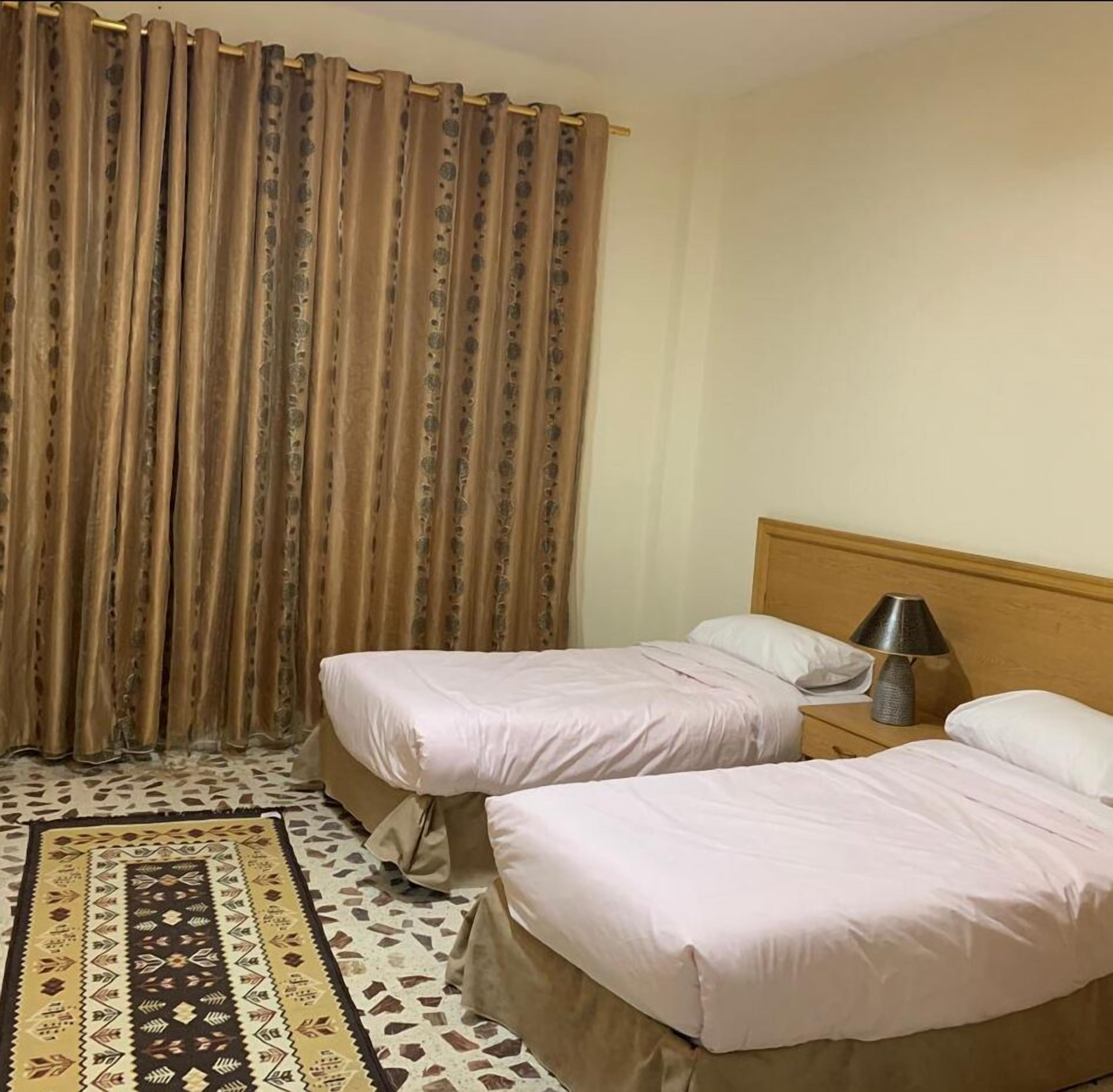 A hotel room with two beds, a patterned rug, a wooden headboard, and a lamp on a bedside table.