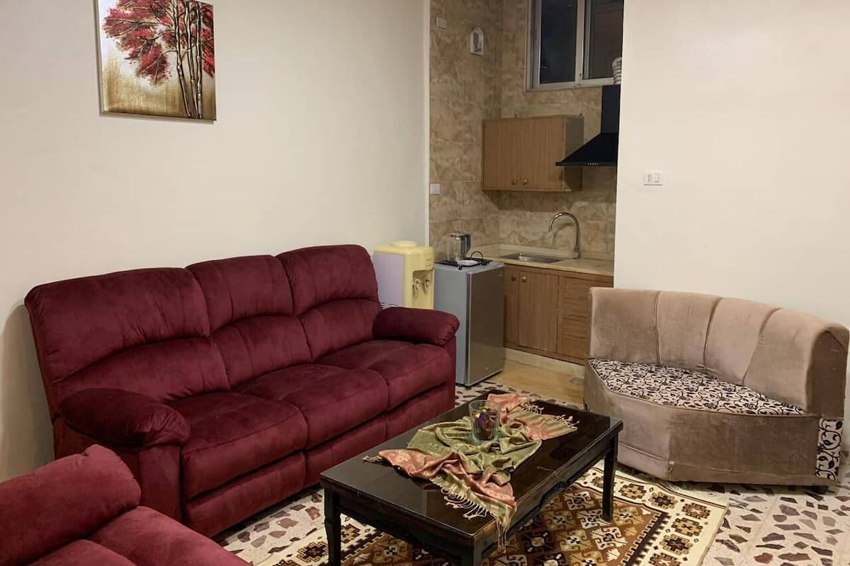 Ein Wohnzimmer mit einem roten Sofa, einem beigen Sofa und einem Couchtisch. Im Hintergrund befindet sich eine Küche mit Holzschränken und einem Fenster.