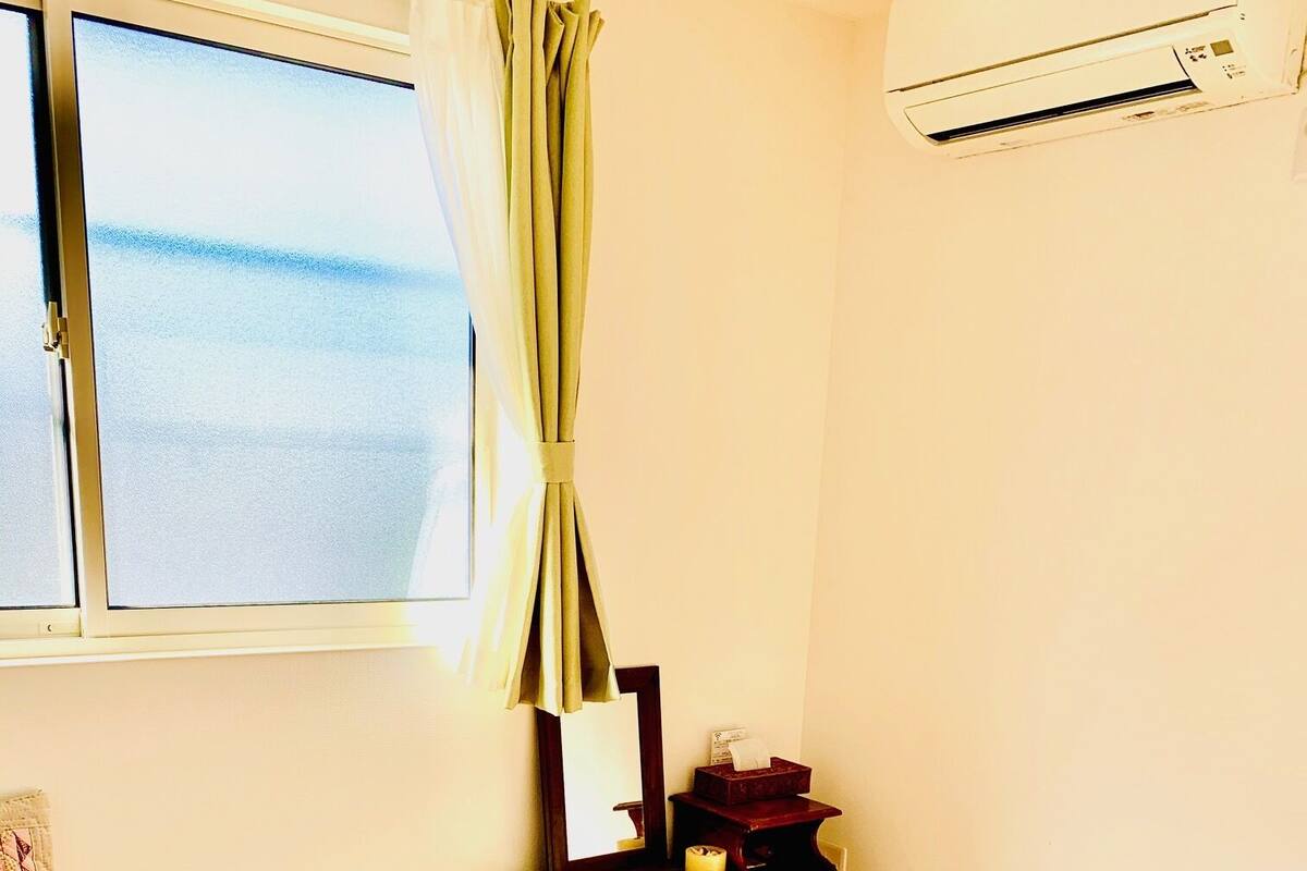 A room with a wooden floor, a window with a view, a small table with a mirror, and a white bucket.