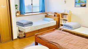 A room with two beds, a small table, a window with curtains, a bookshelf, and an air conditioning unit. - Guesthouse IKETA (Niijima)