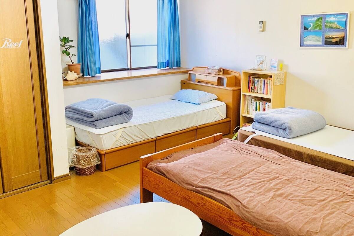 A compact hotel room with two beds, a small table, a bookshelf, and a window with curtains.