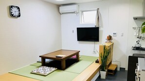 A compact room with a tatami mat bed, a small wooden table, a TV, a kitchenette, and a clock on the wall. - Guesthouse IKETA (Niijima)