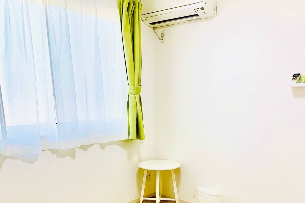 A room with a beige carpet, a white chair, a small white table, a green curtain, and a wall-mounted air conditioner.