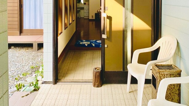 A corridor leading to a room with a sliding door, a small outdoor seating area with chairs, and a visible kitchen area in the background. - Guesthouse IKETA (Niijima)