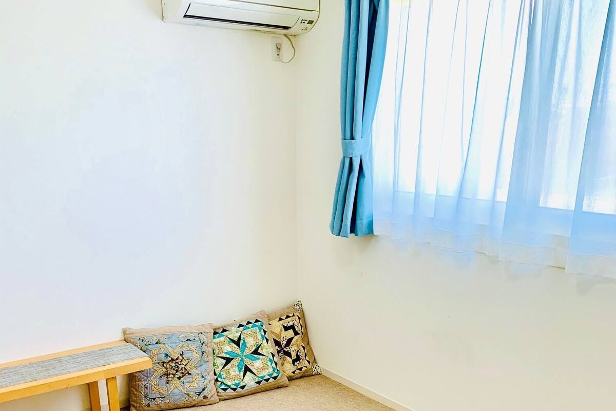 A room with beige carpet, a wooden bench, a patterned cushion, and a window with blue curtains.