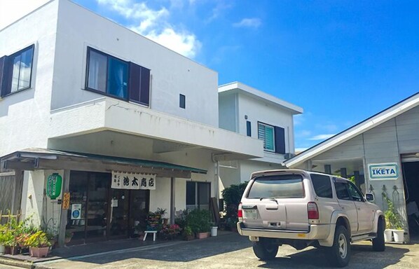 A two-story building with a white facade, a parked SUV, and a visible storefront with signage. - Guesthouse IKETA (Niijima)