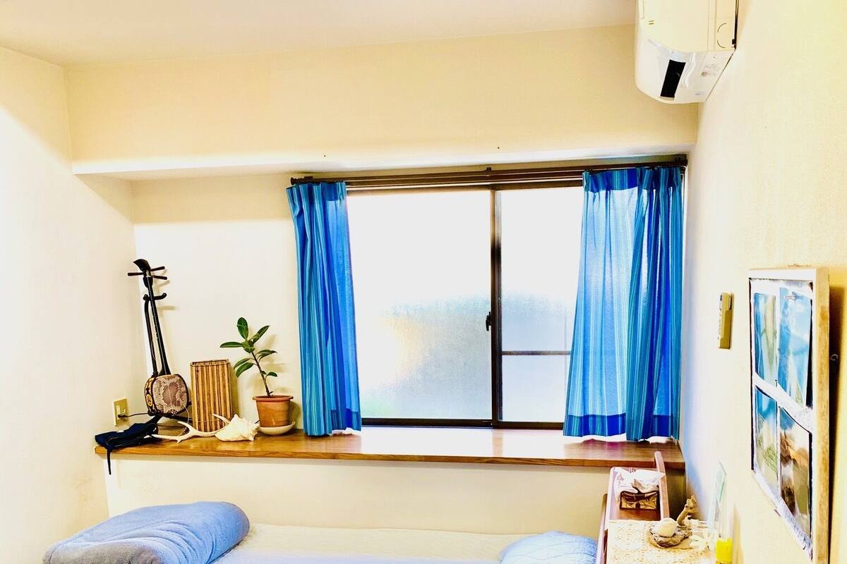 A small, well-lit room with a single bed, a small table, a window with curtains, and a wall-mounted air conditioner.