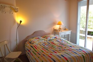 A bedroom with a bed, a chair, a nightstand, a lamp, and a window with a view of greenery.