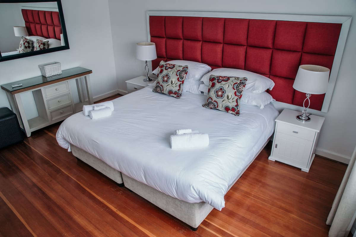 Una cama bien tendida con sábanas blancas y almohadones decorativos, una mesita de noche con lámpara, y piso de madera.