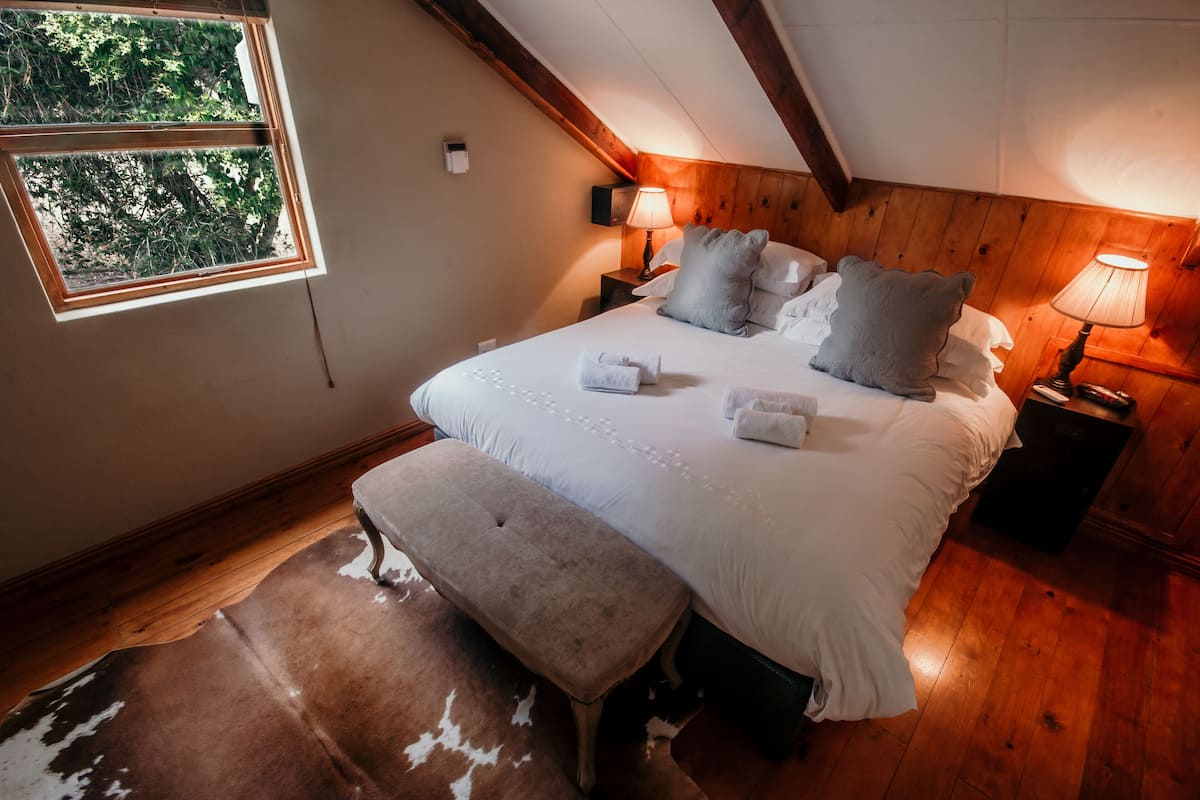 Un dormitorio con una cama grande, cabecera de madera, dos lámparas de noche, una ventana con vistas a árboles y una alfombra de cuero de vaca.