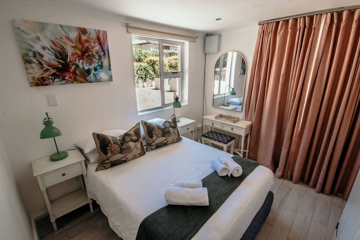 Una cama bien hecha con almohadas, una lámpara de escritorio verde, una mesita de noche, un espejo y una ventana con cortinas.