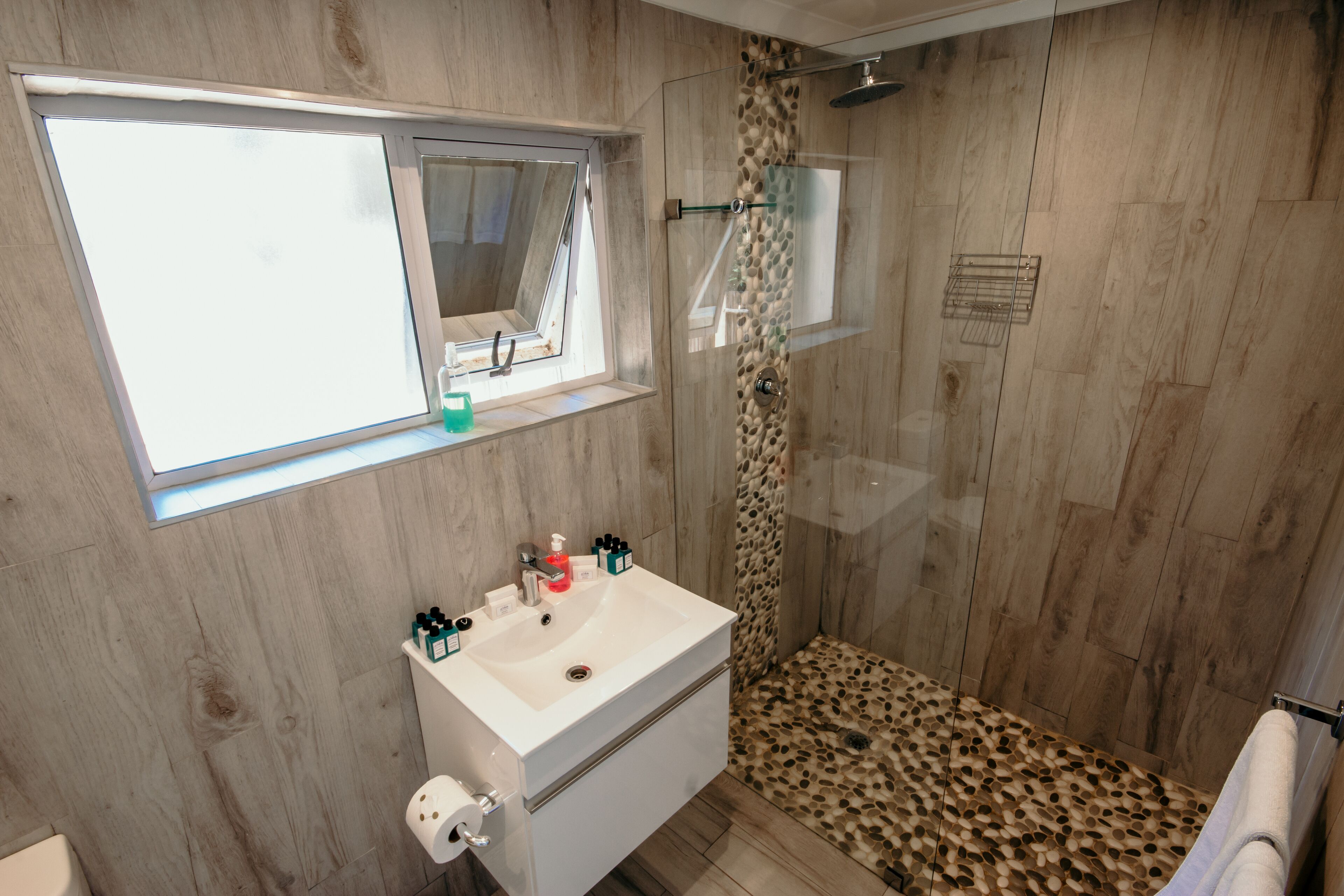 Een moderne badkamer met een houten vloer, een witte wastafel met toiletartikelen, een douche met een luipaardprint vloer en een raam boven de wastafel.