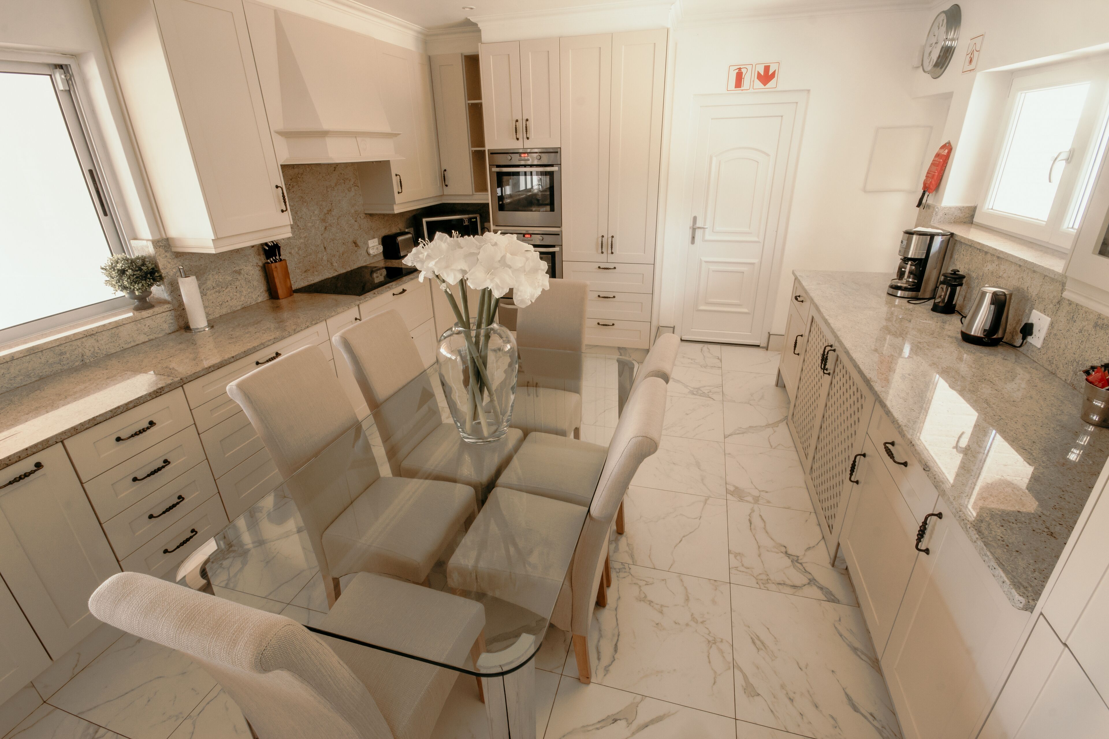 A modern kitchen with white cabinetry, a marble countertop, and a dining area with a glass table and chairs.