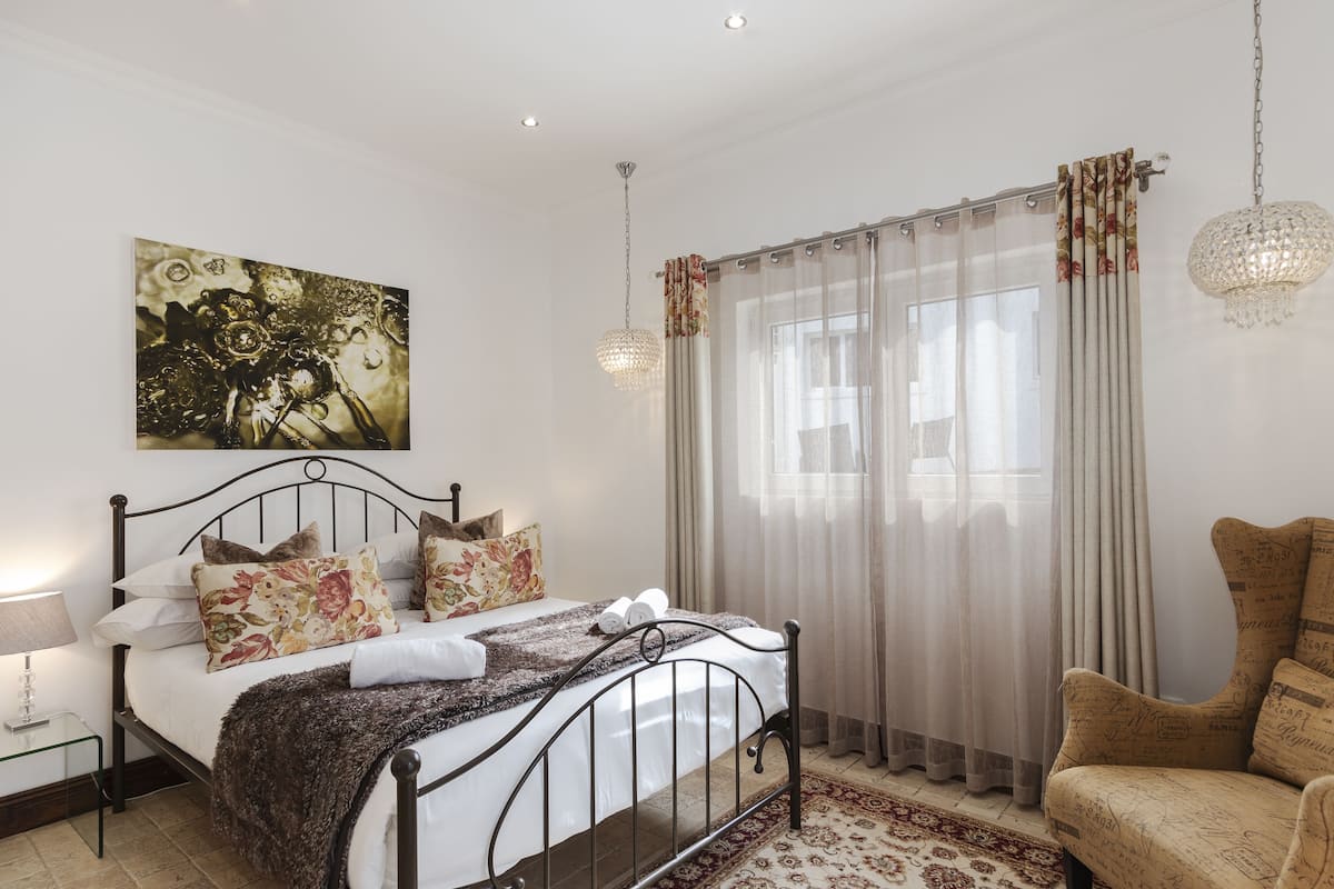 Un dormitorio con cama, mesita de noche, silla, un cuadro y ventana con cortinas.