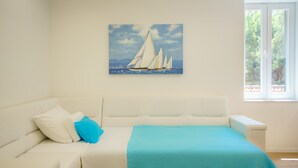 Un sofá blanco con una manta azul y almohadas, un cuadro de veleros en la pared, y una ventana que da a vistas de vegetación.