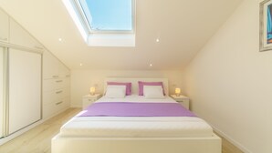 Un dormitorio con tragaluz, una cama con ropa de cama blanca y morada, una mesita de noche con lámpara y un armario.