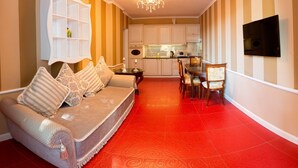 A living room with a sofa, a dining table with chairs, a kitchen area, and a TV mounted on the wall.