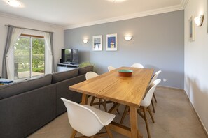 A dining area with a wooden table and white chairs, a grey sofa, and a television on a stand.