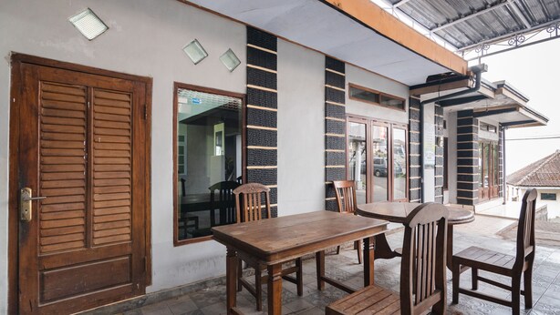 A covered outdoor seating area with a wooden table and chairs, a large wooden door, and a window with a view of the interior. - OYO 1765 Herman Homestay (Wonokitri)