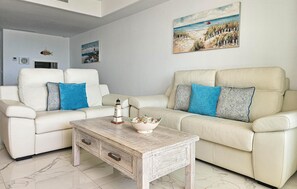 Uma sala de estar com dois sofás brancos, uma mesa de centro e uma pintura de uma praia.