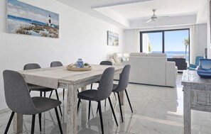 Uma sala de jantar com uma mesa de madeira, cadeiras cinzas, um sofá branco e vista para o oceano através de grandes janelas.