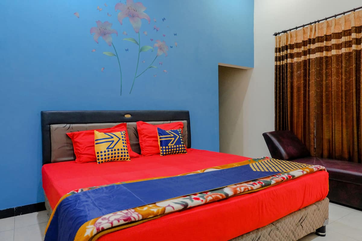Una cama con una colcha roja y varias almohadas, una pared con una obra de arte floral, y una cortina.