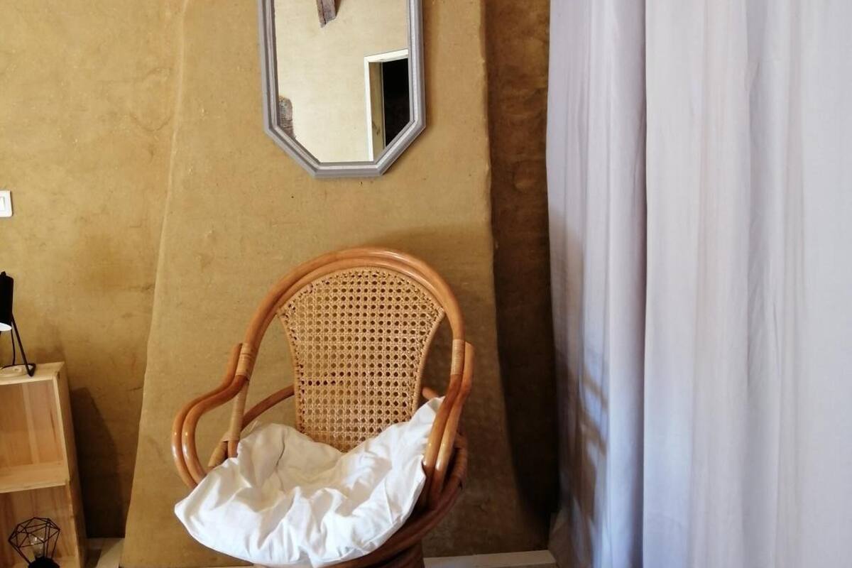 A wicker chair with a cushion, a small round table, a mirror on the wall, and a curtain.