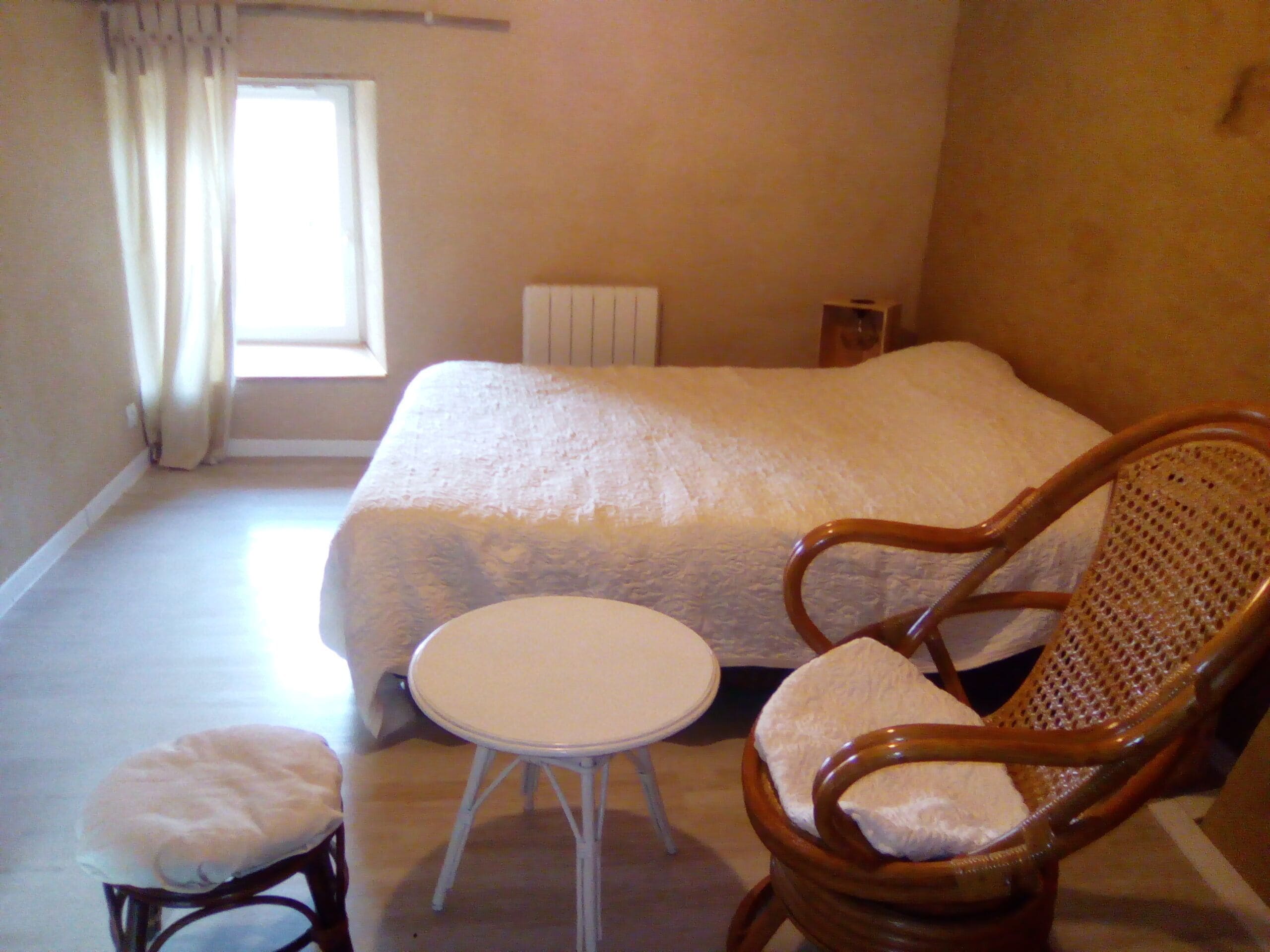 A bedroom with a large bed, a wooden chair with a cushion, a small round table, and a window with curtains.