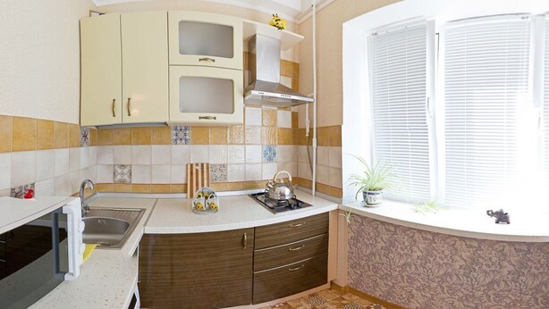 A compact kitchen with a sink, stove, and cabinets. There's a window with blinds and a small plant on the windowsill. - Apart Kiev Starokievskiy pereulok 5-1 (Kyiv)