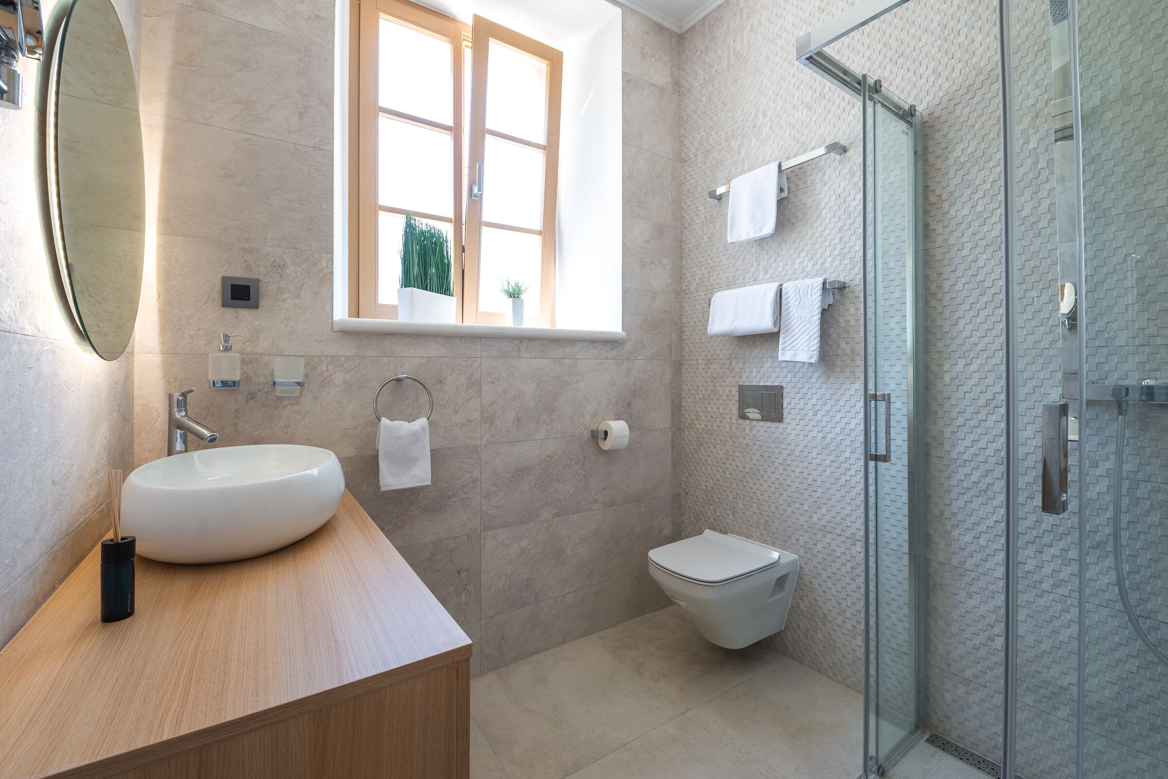 A modern bathroom with a large oval sink, a toilet, a glass shower enclosure, and a window with a view of greenery.