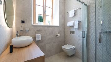 A modern bathroom with a large oval sink, a toilet, a glass shower enclosure, and a window with a view of greenery.