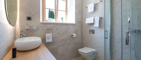 A modern bathroom with a large oval sink, a toilet, a glass shower enclosure, and a window with a view of greenery.