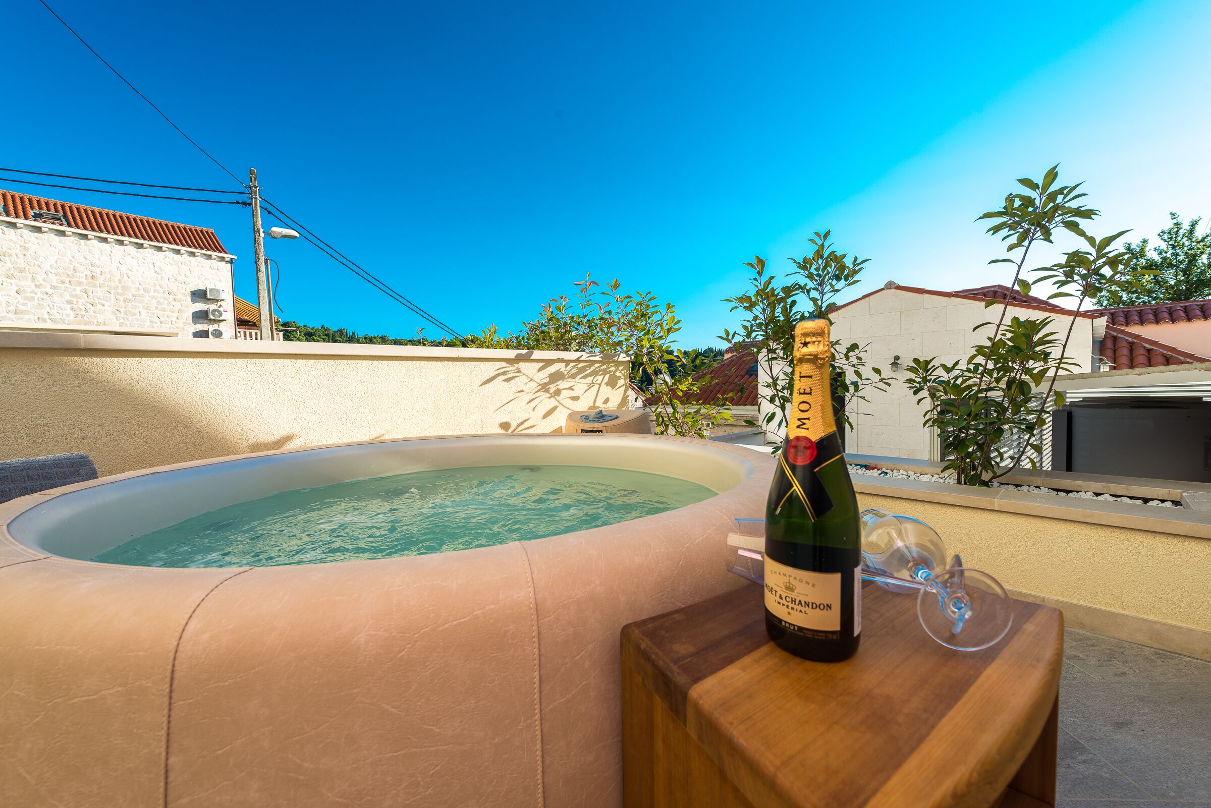 A hot tub with a bottle of champagne and a glass on a wooden table, surrounded by a beige wall and plants.