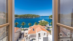 Vista desde una ventana hacia una zona costera con mar azul cristalino, una pequeña isla y edificios con tejados rojos.