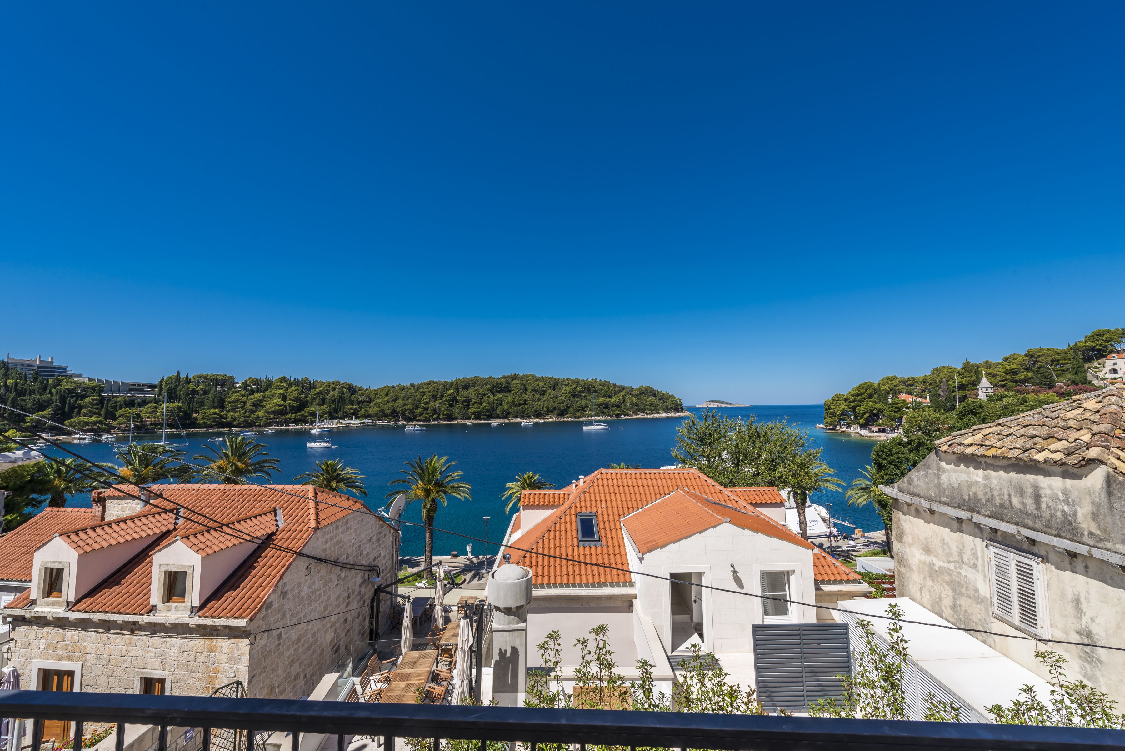 A coastal town with terracotta-roofed buildings, a marina with boats, and a clear blue sky.