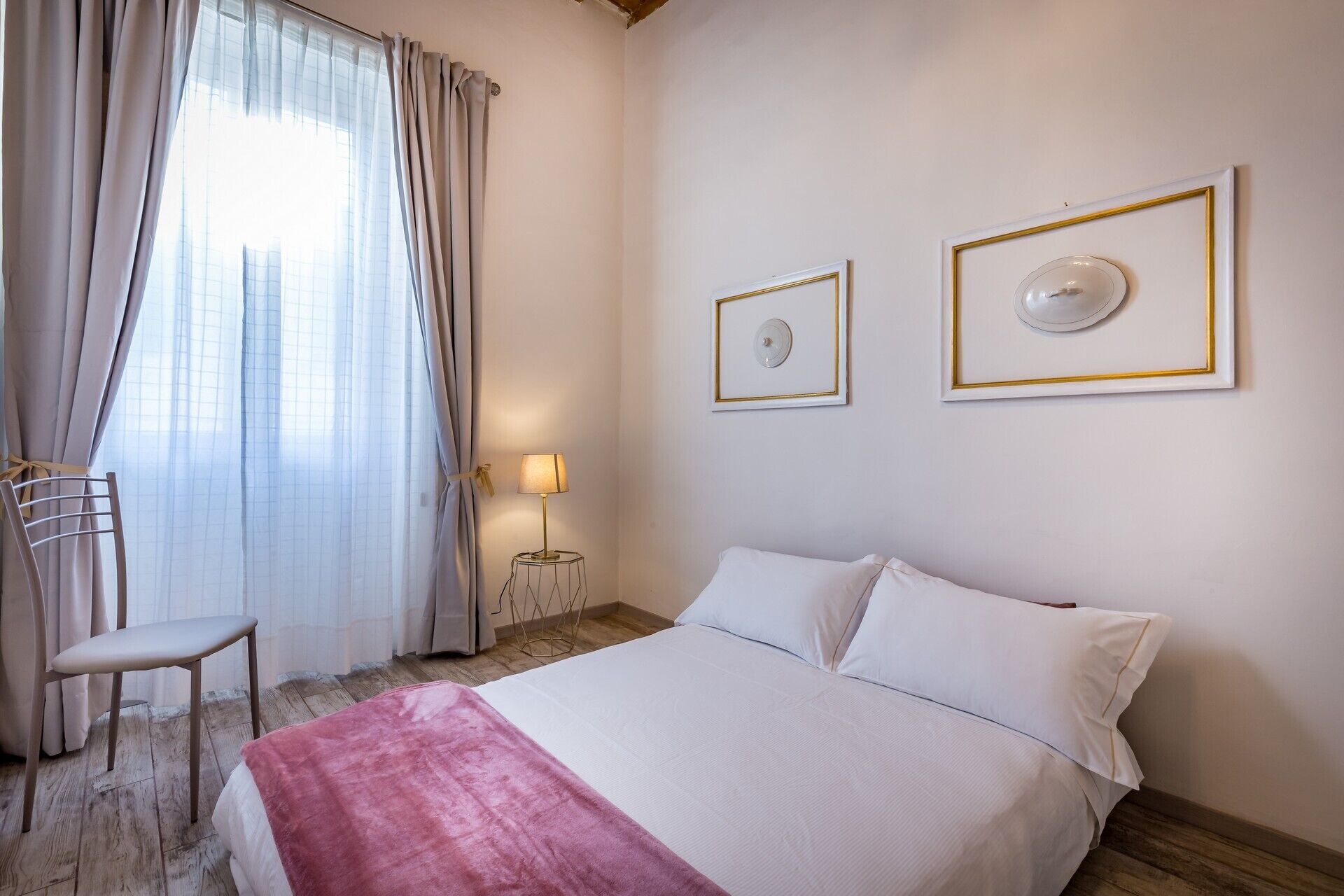 A hotel room with a bed, a chair, a lamp, and two framed pictures on the wall.