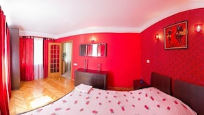 A bedroom with a red wall, a wooden floor, a bed with a floral bedspread, a dresser, a mirror, and a door leading to another room. - Apart Kiev Starokievskiy pereulok 5-2 (Kyiv)