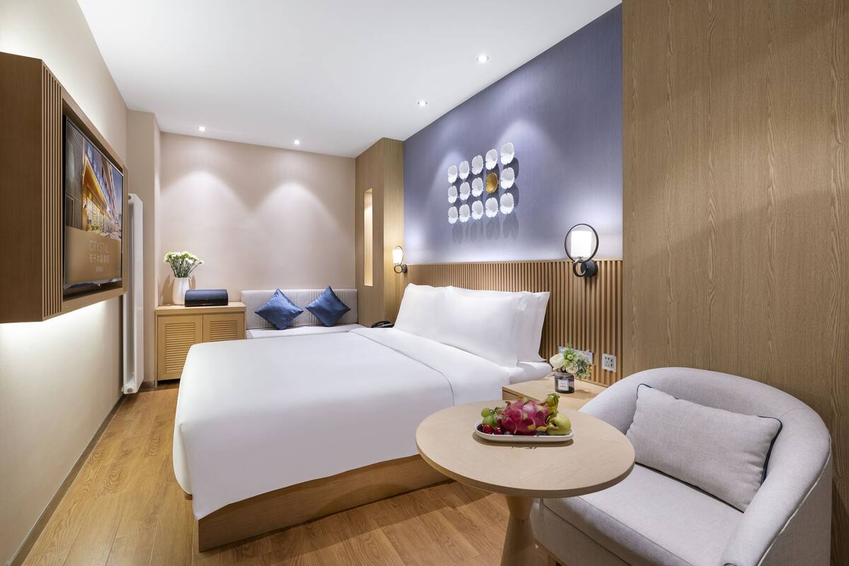 Ein modernes Hotelzimmer mit einem großen Bett, einem runden Tisch mit Obst, einer Couch und einer Wandleuchte.