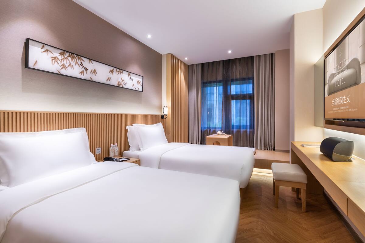 Ein Hotelzimmer mit zwei Betten, einem hölzernen Kopfteil, einem Nachttisch mit Telefon, einem kleinen Tisch mit Lampe, einem großen Fenster mit Vorhängen und einem an der Wand montierten Fernseher.
