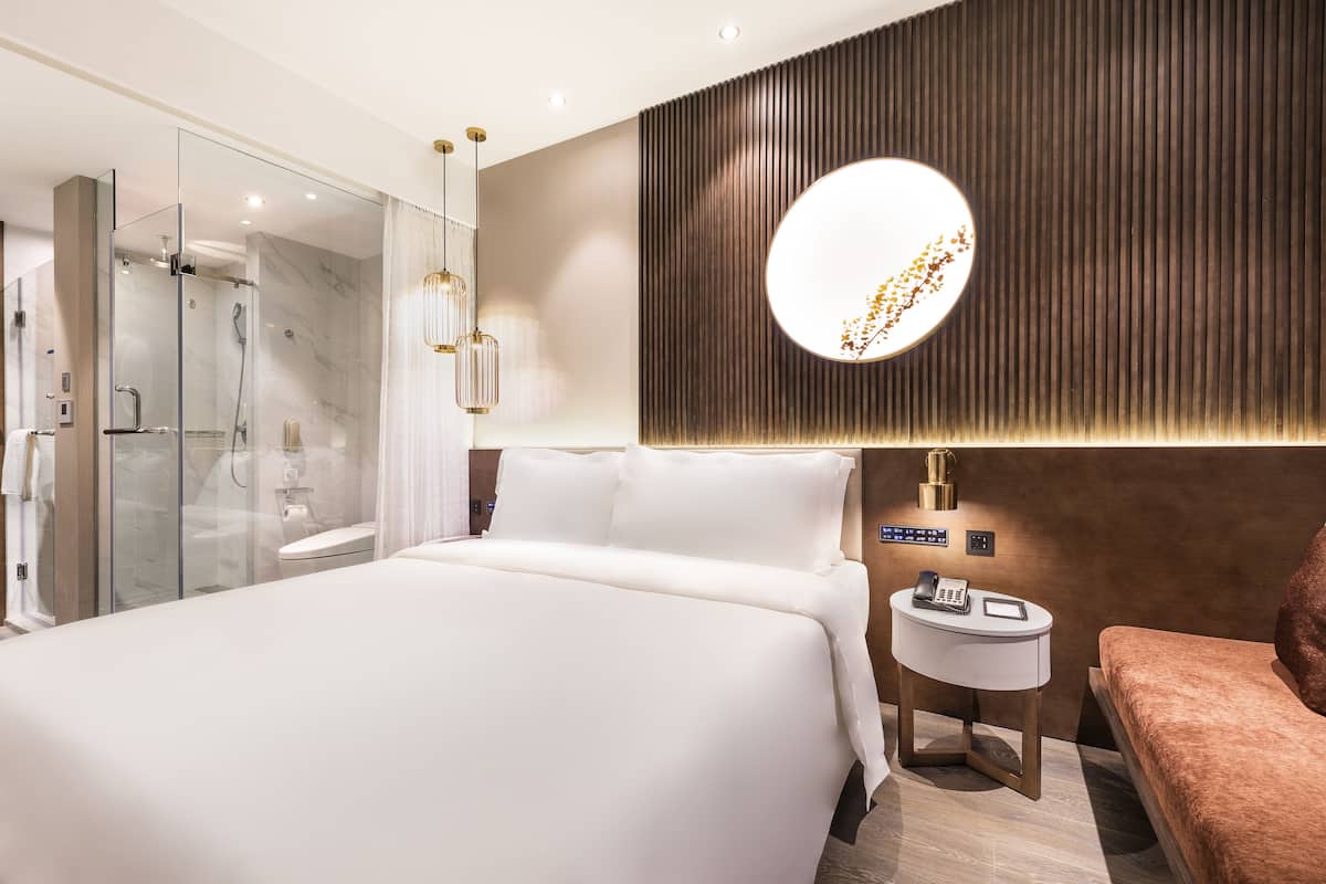 Ein modernes Hotelzimmer mit einem großen Bett, einer Glaskabine für die Dusche, einem runden Spiegel und einem kleinen Nachttisch mit Telefon.