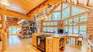Una cabaña de troncos espaciosa con una gran mesa de comedor de madera, una cocina con estufa y una chimenea de piedra.