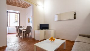 Een moderne woonkamer met een witte bank, een eethoek met stoelen en een tafel, een televisie op een witte kast, en een schaal fruit op een salontafel.