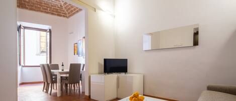 A modern living room with a white sofa, a dining area with chairs and a table, a TV on a white cabinet, and a bowl of fruit on a coffee table.