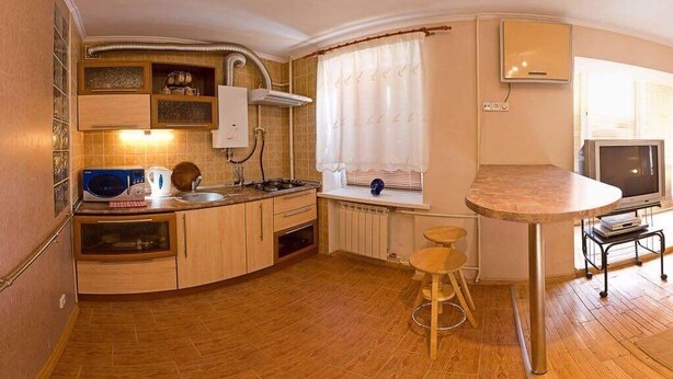 Ett kompakt kök med mikrovågsugn, spis och diskho, en liten matplats med pallar och en tv.