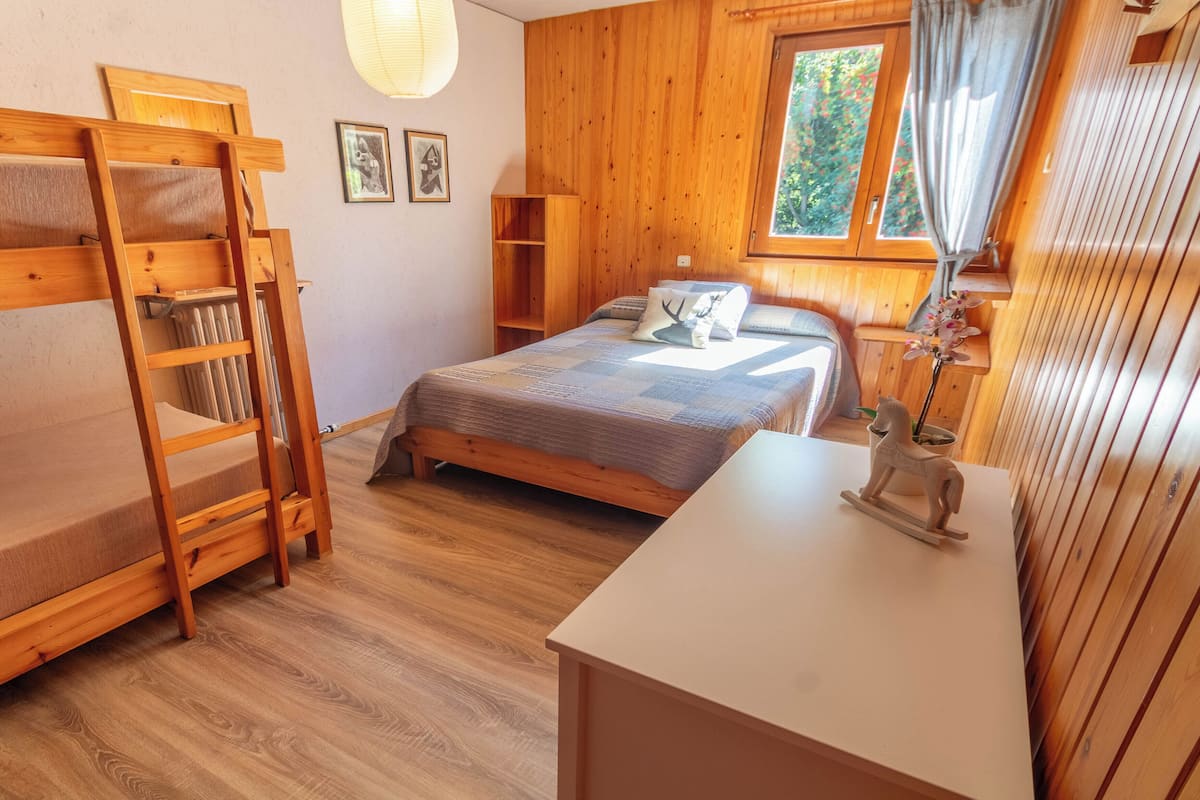 Ein gemütliches Zimmer mit Etagenbett, Einzelbett, einem Holztisch und einem Fenster mit Blick auf Bäume.